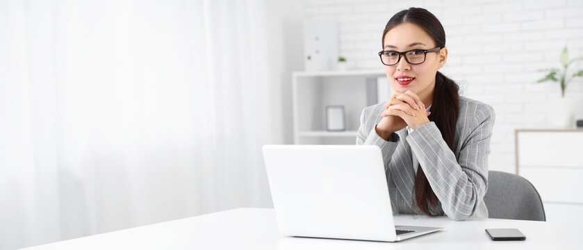 Young Asian businesswoman with modern laptop and mobile phone sitting at her workspace in office