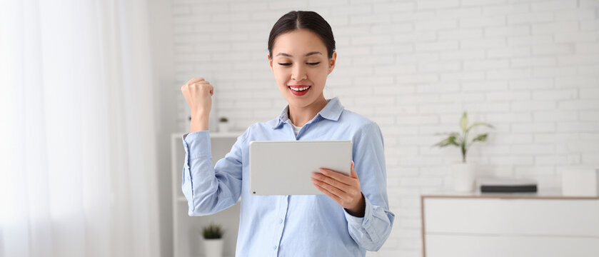 Happy young Asian businesswoman with modern tablet computer celebrating success in office