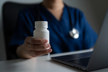 Doctor in blue scrubs holding white medicine bottle near laptop, representing telemedicine, online prescription, remote healthcare consultation, and modern digital medical service.