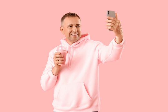 Mature man with glass of fruit smoothie and mobile phone taking selfie on pink background