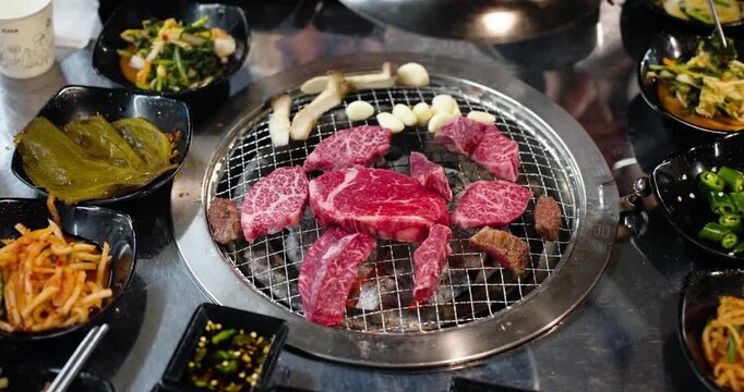 Top-down view of beautifully marbled raw Hanu beef slices, mushrooms, and garlic cooking on a circular charcoal grill, surrounded by an assortment of delicious Korean banchan side dishes