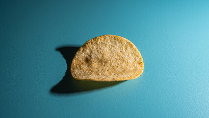 Naklejka premium A single, perfectly shaped, seasoned potato chip rests on a vibrant teal blue surface, casting a long, dramatic shadow under focused studio lighting, emphasizing its texture.