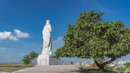 A huge statue of Christ made of white Carrara marble on a hilltop against the blue sky. Silhouettes of people on the observation deck.   City in the distance. A green tree in the foreground. Havana.  