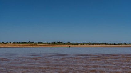 A calm red-brown African river. There is a silhouette of a man on the sandy shore. Green vegetation in the distance. Clear blue sky. Copy space. Madagascar. Tsiribihina   river.