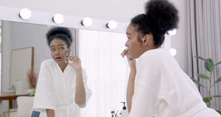 African teenage girl curling lashes using eyelash curler while smiling in mirror, preparing face for makeup during self care routine in bright bedroom with cozy and modern interior style - Powered by Adobe