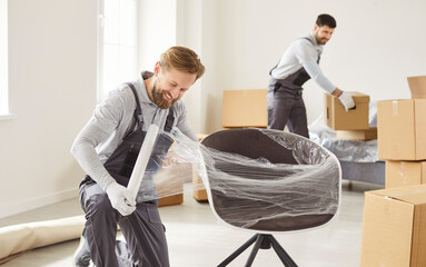 Movers team at work, smiling men wrapping office chair furniture in plastic cover to protect,...