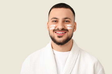 Young man in bathrobe with cream smears on cheeks against white background
