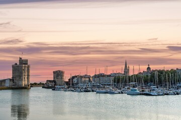 Obraz premium Witness the breathtaking sunset over La Rochelle, France. Historic towers guard the harbor as boats gently sway. The sky is a canvas of pink and orange, creating a serene atmosphere.