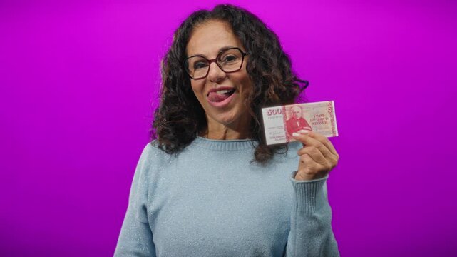 Middle-aged woman holding icelandic krona smiling against vibrant purple background.