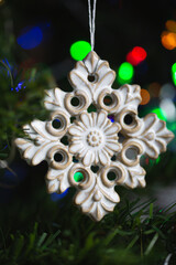 Ceramic toy in the form of a snowflake hanging on a Christmas tree with lights from a garland in the background.