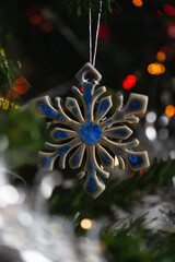 Ceramic toy in the form of a snowflake hanging on a Christmas tree with lights from a garland in the background.