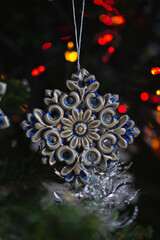 Ceramic toy in the form of a snowflake hanging on a Christmas tree with lights from a garland in the background.