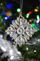 Ceramic toy in the form of a snowflake hanging on a Christmas tree with lights from a garland in the background.