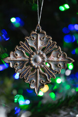 Ceramic toy in the form of a snowflake hanging on a Christmas tree with lights from a garland in the background.