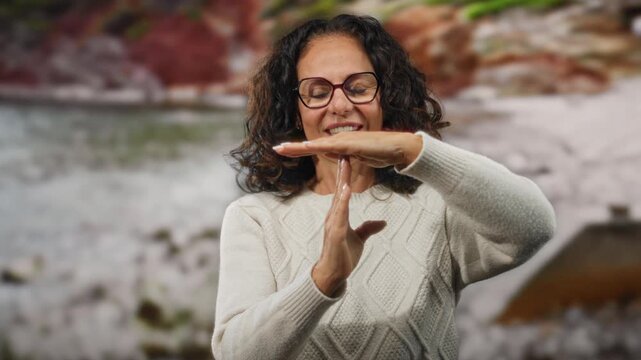 Woman with curly hair making timeout gesture at the seaside, wearing glasses and a sweater at the beach, giving a mature expression of calm serenity and relaxation outdoors.