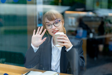 Woman enjoying a coffee while working at a cafe and waving to the camera during a sunny afternoon...