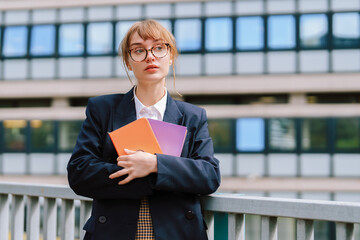 Young woman in glasses holds colorful notebooks while standing near modern building in early afternoon light