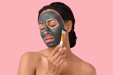 Pretty young African-American woman with brush and activated charcoal mask on her face against pink background