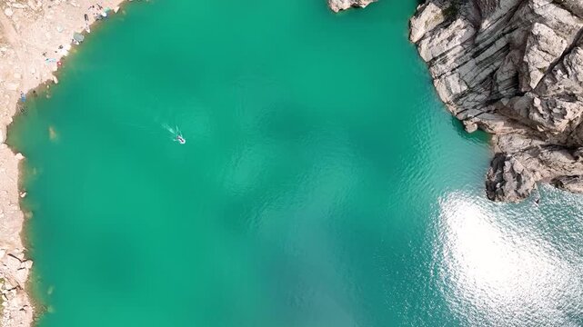 Direct aerial view of the brilliant turquoise water of Kel-Suu Lake showing a small boat, sun glare, and a rocky shoreline with a few people and tents