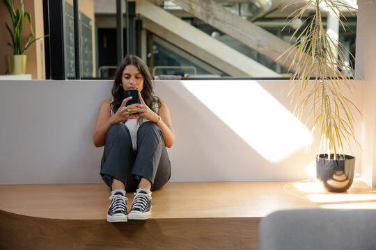 Woman sitting on a ledge indoors, scrolling on her smartphone calmly