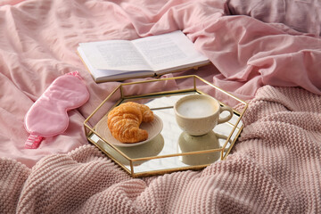 Cup of coffee with croissant, book and sleep mask on pink bed sheets