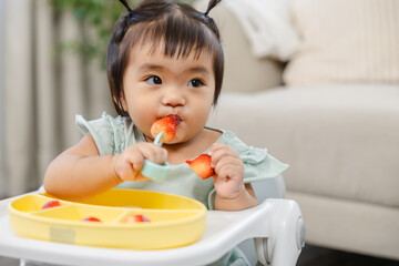 Happy toddler in high chair tastes strawberry and soft bites, practicing hand-to-mouth coordination and building eating habits; pediatric nutrition, growth milestones, child development, family care
