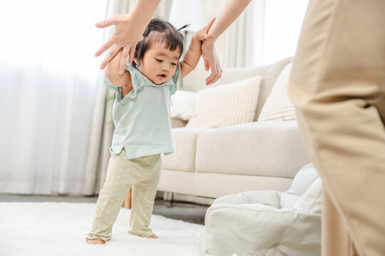 One-year-old baby learning to walk with parent support at home, first step moment showing growth, balance, and family bonding, first walking practice, showing curiosity, healthy childhood milestone - Powered by Adobe