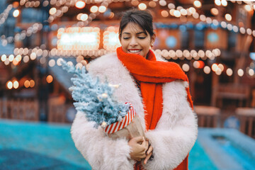 Smiling woman in cozy white coat holds small blue Christmas tree with red and white decoration at festive outdoor market