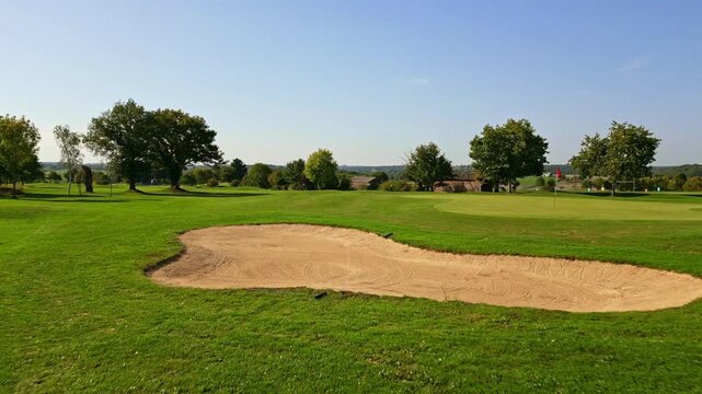 Low-altitude drone shot showing a sand bunker, golf flag, trimmed green grass, and surrounding trees under clear sky.