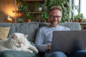 A happy caucasian man with glasses working on his laptop on a comfortable couch next to his sleeping fluffy cat in a cozy apartment
