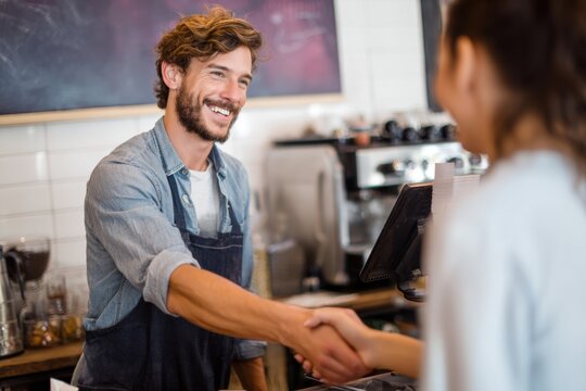 A smiling young caucasian man wearing an apron shakes hands with a customer while working at a coffee shop counter