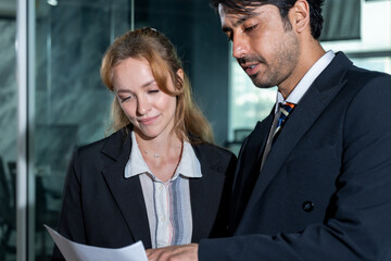Asian businessman and caucasian businesswoman reviewing business document together in hallway of modern office, discussing work progress and team performance in professional setting