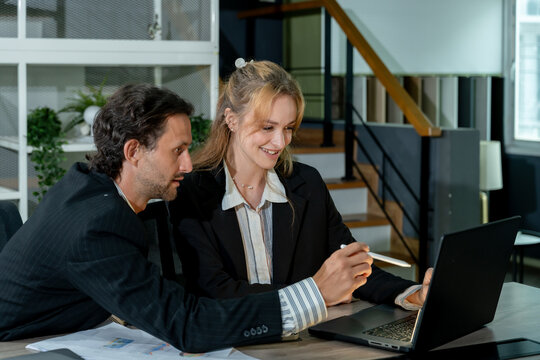 Caucasian businessman and caucasian businesswoman smiling while working together on laptop in modern office, discussing company data and digital marketing strategy for business development