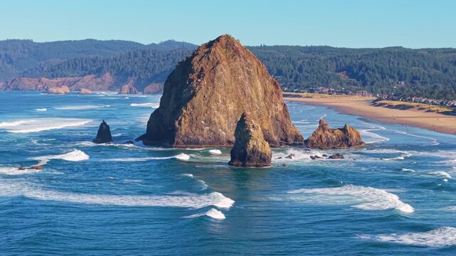 A breathtaking high-angle video capturing the iconic Haystack Rock sea stack, the expansive sandy beach, and the Pacific Ocean coastline under a clear, bright, sunny sky at Cannon Beach, Oregon