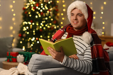 Young man in Santa hat reading book at home on Christmas eve