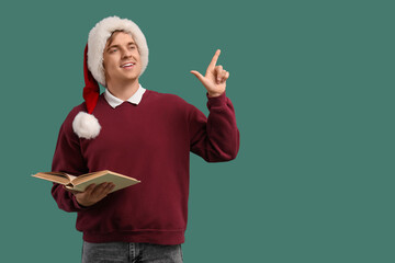 Young man in Santa hat and with book pointing at something on green background