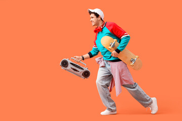 Happy young man with headphones, skateboard and tape-recorder running on orange background