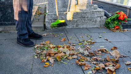 Person sweeping autumn leaves from cemetery walkway near grave, preparation for All Saints Day and caring for memorial resting place of loved ones