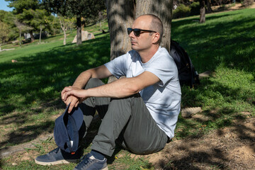 Middle  aged man in sunglasses holding his cap is chilling at a park on a sunny day.
