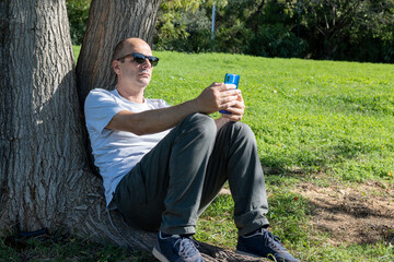 A middle aged Caucasian man wearing sunglasses is leaning to a tree, relaxing at a park watching his smartphone.