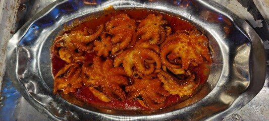 Cooked baby octopuses in spicy red sauce served on a metal plate — a rich seafood dish full of flavor and Mediterranean aroma.