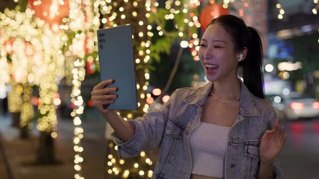 Portrait of Asian woman talks on video call via digital tablet in night megapolis. Happy Korean lady waves hand Hello to friend online on downtown street slow motion. Internet chat