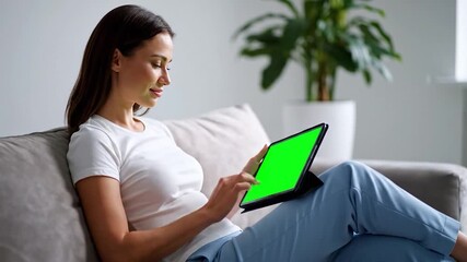A woman is on a grey sofa, using a tablet with a green screen, indoors near plants - Powered by Adobe