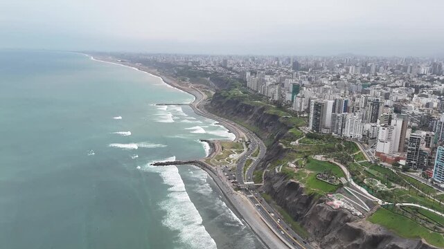 Camera flies along Miraflores boardwalk capturing heavy waves hitting brick structures, traffic below cliffs, green parks, and high-rise buildings in Lima, Peru