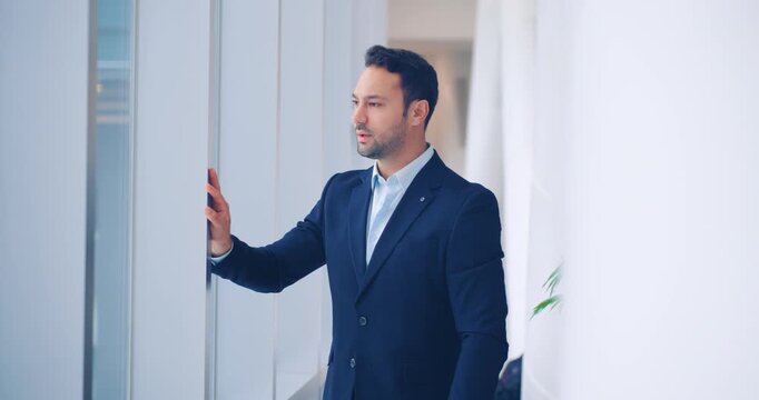 Handsome confident European businessman standing composed in a bright minimalist corporate office corridor looking determined checking time on wrist watch and while looking at the window