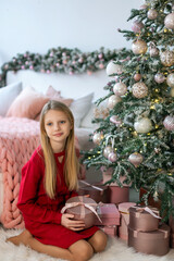 Young girl with Christmas gift near tree. A blonde girl in a red dress holding a round gift box while sitting beside a decorated Christmas tree in a cozy holiday room.