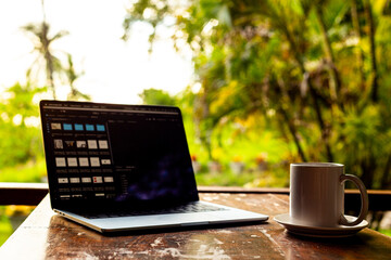 Laptop and cup on wooden table of terrace with tropical forest view at sunset, remote work digital nomad.
