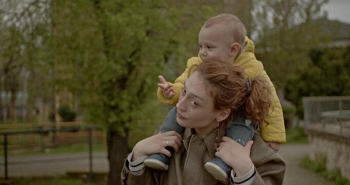 A mother carries her toddler on her shoulders in a park setting. They point and wave, enjoying their time together outdoors.