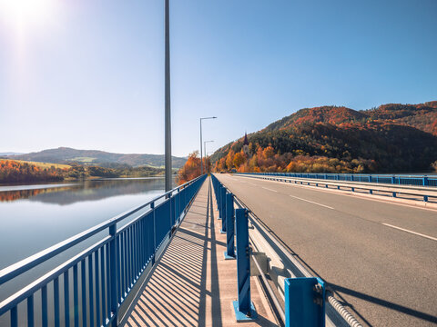 Pedestrian walkway and road bridge in Margecany next to the Ružín reservoir, with calm water and autumn-colored hills in the background under a clear sky.