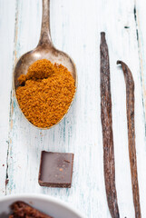 pile of brown sugar on old spoon, vanilla, broken dark chocolate blocks on white wooden table